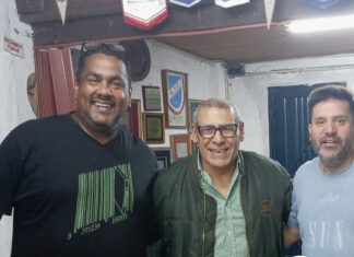 Bien por ellos Group of three smiling men posing for a photo in a room with plaques on the wall; a green sports banner at the bottom reads 'Deportes' and 'Bien por ellos' with additional article text.