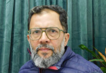 Una mirada crítica al desembarco de 1825 Portrait of a middle-aged man with glasses and a gray beard, wearing a blue puffer jacket, in front of green curtains.