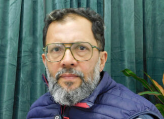 Una mirada crítica al desembarco de 1825 Portrait of a middle-aged man with glasses and a gray beard, wearing a blue puffer jacket, in front of green curtains.