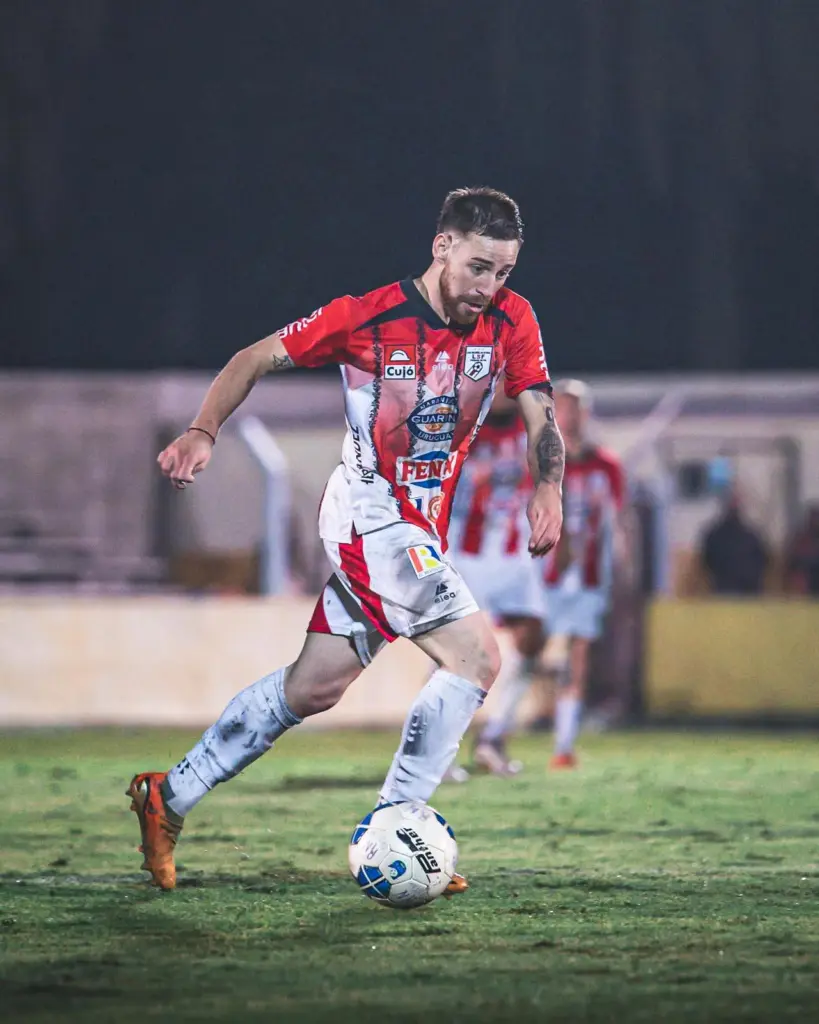 Soccer player in red-and-white kit dribbling a ball on a grass field during a night match.