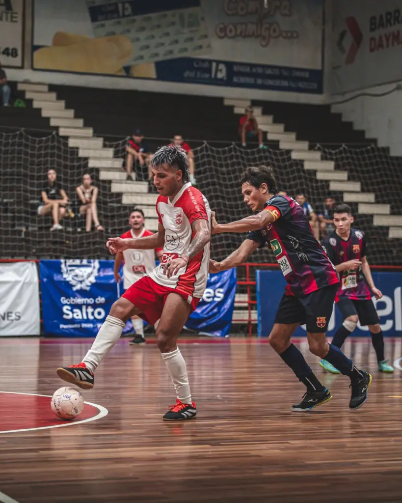 Futsal players compete for the ball on a polished wooden court, white/red player in front and a blue/purple opponent applying pressure with a netted barrier and spectators in the background.