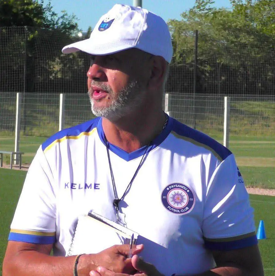 Male soccer coach wearing a white cap and a white jersey with Paysandu crest, holding a clipboard on a practice field.